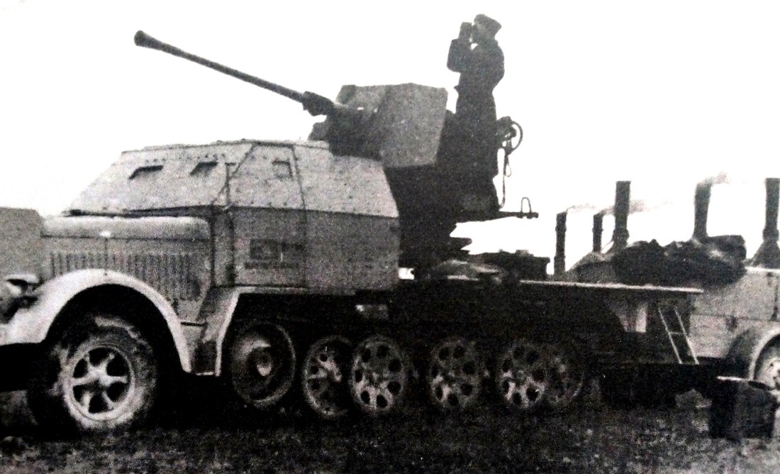 一辆在德国某重点工厂防空阵地上的Sd.Kfz.7/2半履带装甲自行炮车。该炮车已经过改装，在舱室和车前格栅加装了80毫米厚的装甲钢板。并拖挂了Sd.Ah 57弹药车。炮车上一德军正在37毫米Flak 36高射炮炮位上作对空观察。