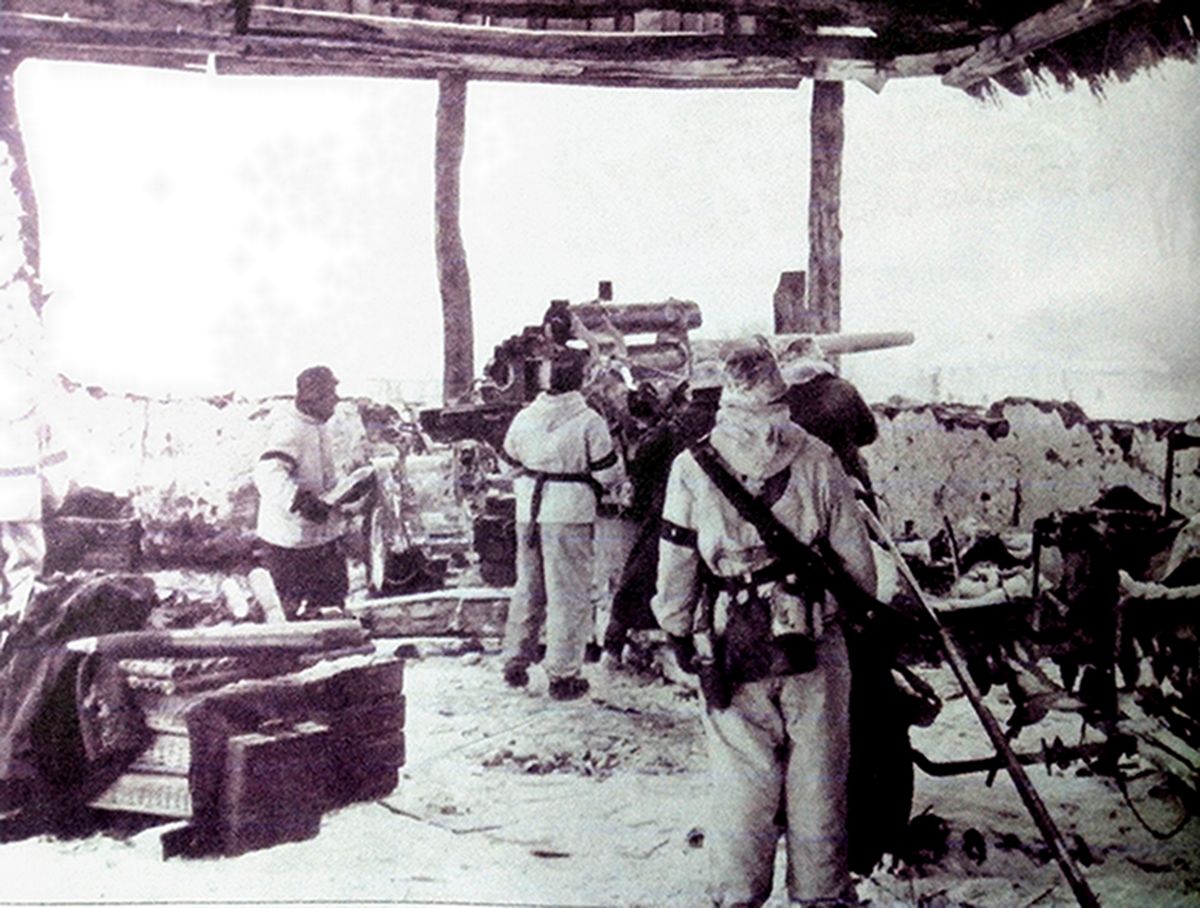 1941年冬天,东线苏联战场,德军将一门88毫米Flak18高射炮的阵地安置在一个废弃的房屋中。为了保证足够的射角,德军将屋子四周的墙壁打掉,只留下支撑屋顶的立木。通过炮管的高度可知,该炮正在与地面目标交火,屋内地上有一堆柳条制的三发弹药箱,除了一弹药手捧着一枚炮弹准备装填外,弹药箱上还放着备用的炮弹。