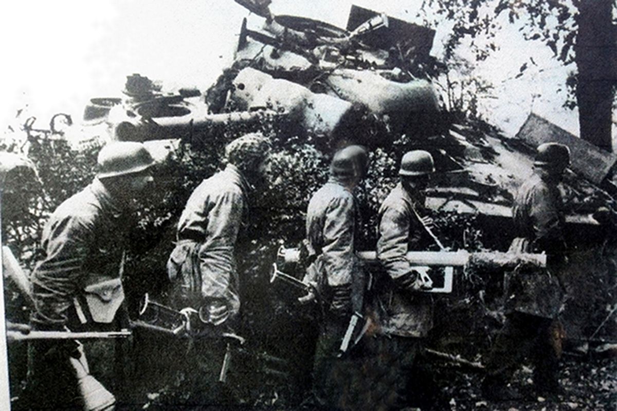 在1944年夏末,西线法国战场,手持「坦克杀手」反坦克火箭筒和「铁拳」反坦克榴弹发射器的德军反坦克排士兵在一辆被击毁的英军M4A4型坦克旁经过,边走边警惕地注视着四周的情况。