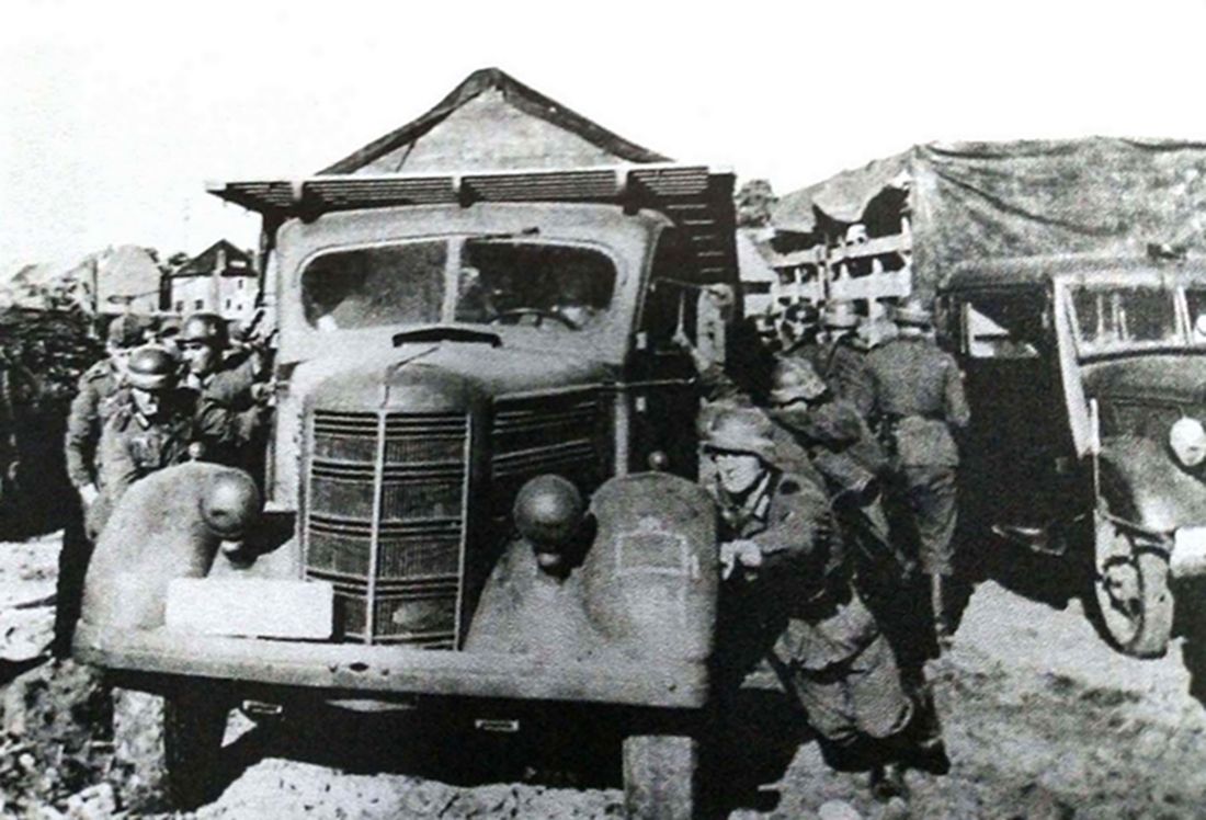 1940年，西线战场比利时地区，一群德军士兵正在用力将发生故障的一辆福特V8-51中型军用卡车推离公路，以免堵住德军物资运输车队前进的道路。