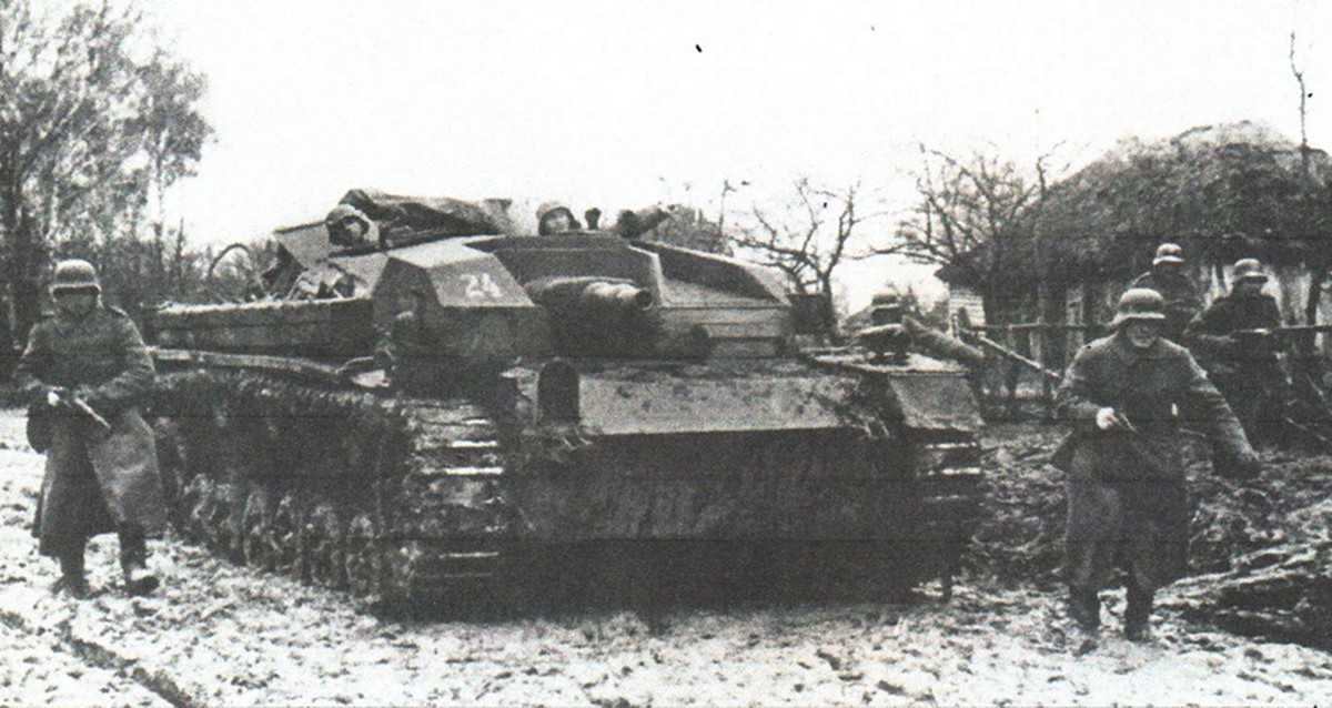1941年秋,东线苏联战场,由于恶劣天气带来的泥泞路况和大雨,图中这辆第192突击炮团的Ⅲ号B型自行突击炮车正与一队步兵配合作进攻姿态,为战地摄影师做摆拍,以达到战事宣传目的。在这辆炮车前甲板上的战术标号下方印有这支部队的浅色「十字骷髅」徽标。而放置在履带装甲上的木条(主要用于应付苏联境内的恶劣路况和反战壕之用)也算是这支部队的特色标志之一。