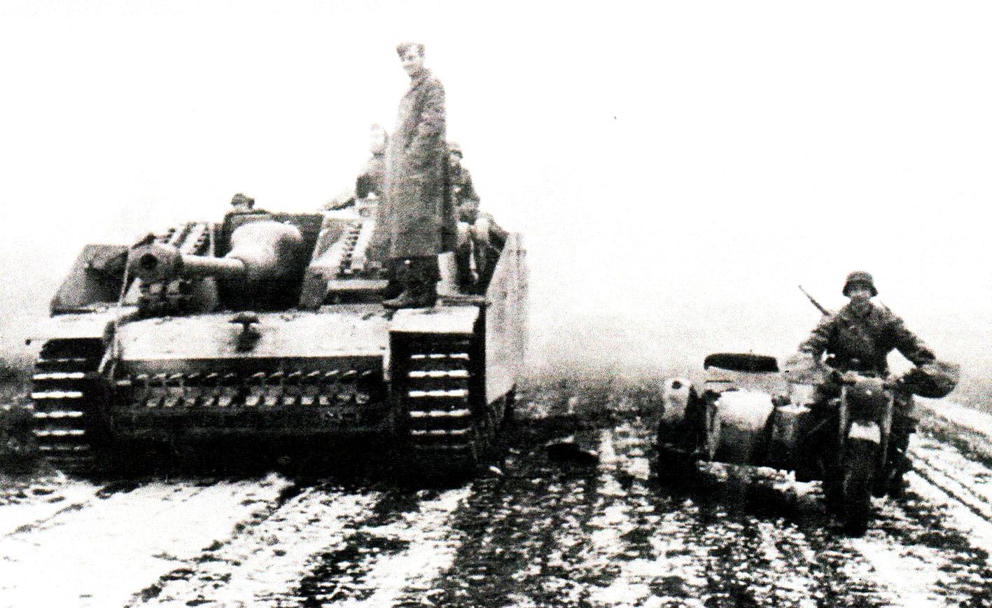 正在等待新命令的一辆StuG ⅢG型突击炮。这辆晚期型的G型车可能是辆新车,状况良好。这可从它炮台前方装甲板上携带的一大段新的备用履带看出来。一个德军通讯兵正驾驶一辆摩托车在车旁驶过。
