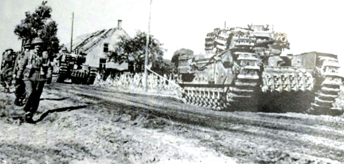 1944年,英军第4近卫步兵营的丘吉尔Ⅵ型步兵坦克行进在荷兰一村庄,跟随坦克行进的是英军萨克福第1步兵营的士兵。注意坦克车体与炮塔外都有附加履带作防护装甲。