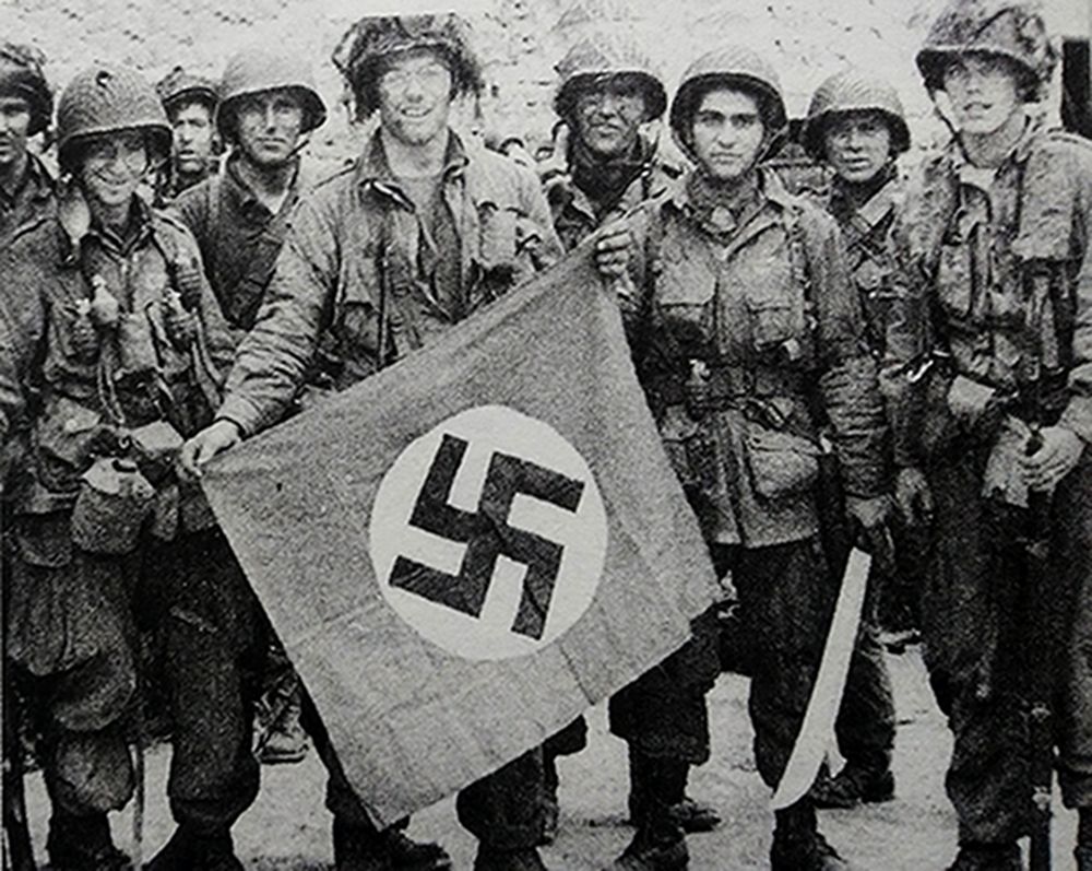 1944年夏天,「诺曼底登陆」期间,西线法国战场诺曼底地区,空降的美军伞兵展示其缴获的一面德军纳粹党旗,站在旗帜左侧的那名士兵脖子上足足挂着4枚MK2手榴弹。