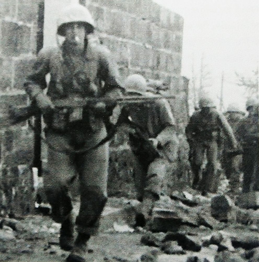 1945年，「冲绳岛战役」期间，太平洋战场，美军一名手持M1918A2「勃朗宁」自动步枪的军士带领战斗小队穿行在废墟中。