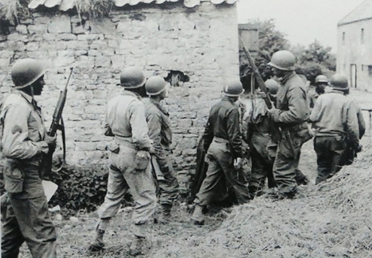 1944年6月，西线德国战场，美军一队士兵包围一个农舍，搜索德军狙击手，排在最后面的一名黑人士兵手持M1卡宾枪。