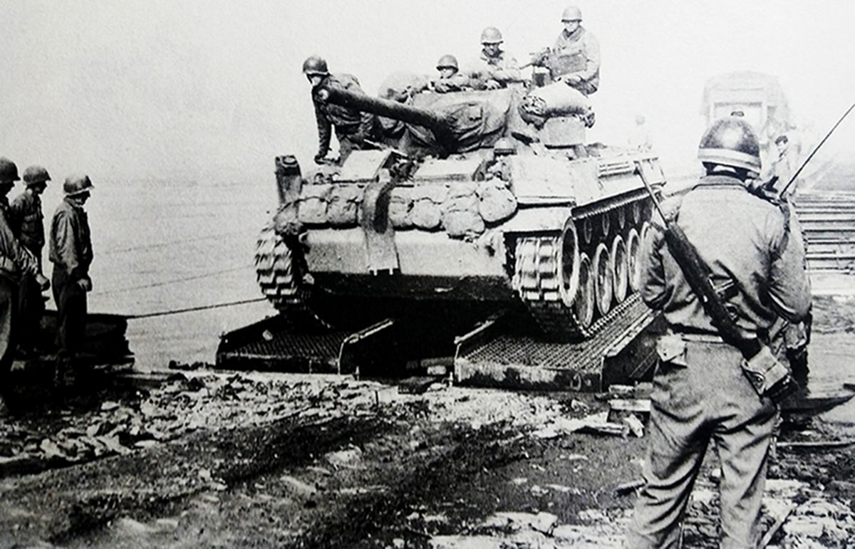 1945年初，西线德国战场，美国巴顿的第3集团军第4装甲师穿越法兰克福挺进威瑟河地区，一辆M18坦克歼击车正在通过摩泽尔河上的临时浮桥，一名负责交通管理的美军宪兵身背M1卡宾枪在桥边值守。