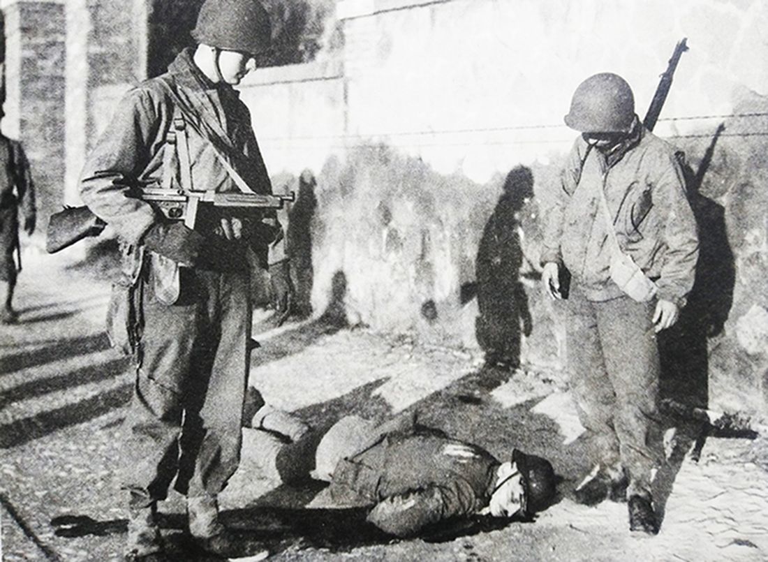 1944至1945年间,欧洲西线战场上,两名美军士兵在一条攻下的城镇街道上注视着一名被击毙的德军士兵,左边的美军士兵肩上挂着一支M1A1汤普森冲锋枪。