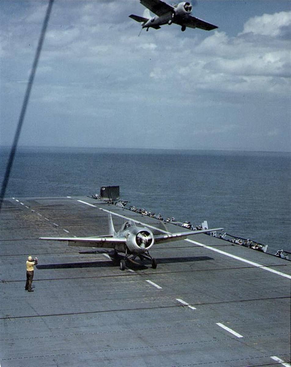 1944年5月8日,切萨皮克湾训练海域,一架通用汽车 FM-2「野猫」战斗机准备从「充电者」号(CVE-30)起飞;上方另一架 FM-2 已放下着舰钩通场,因甲板被占而收到复飞信号。两机均涂大西洋战区浅色迷彩。此时该舰正担负新飞行员的舰上资格训练。
