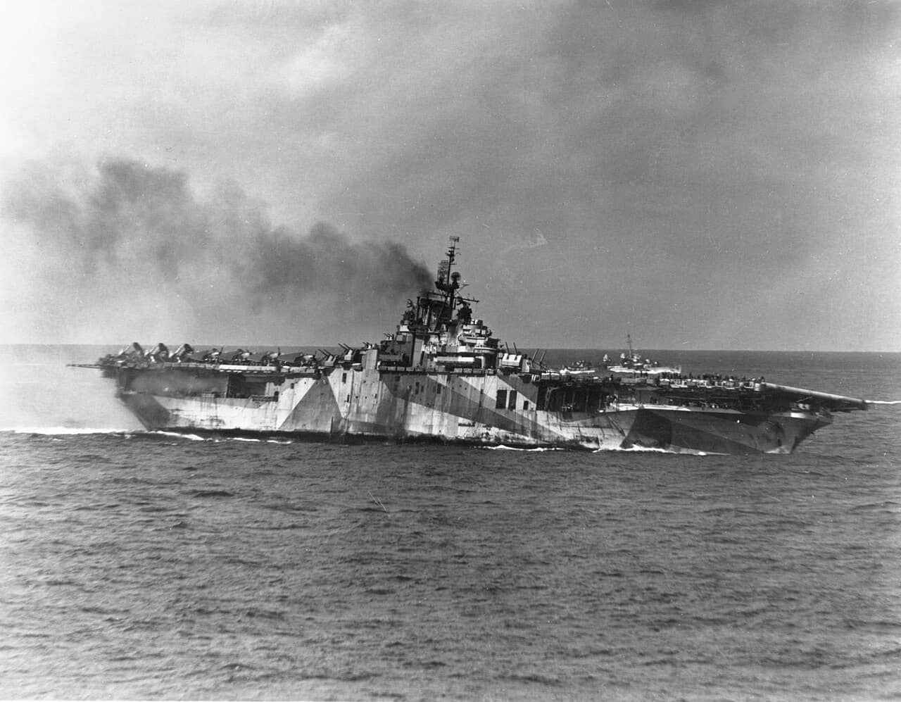 1945年1月21日,美国海军埃塞克斯级提康德罗加号航空母舰(USS Ticonderoga,CV-14)遭到「神风」特攻队袭击后舰体向左舷倾斜。当时,共有四架自杀式飞机撞击了该舰。注:其舰身采用了「33号伪装方案10A式样」的迷彩涂装,背景中还有一艘弗莱彻级(Fletcher-class)驱逐舰。
