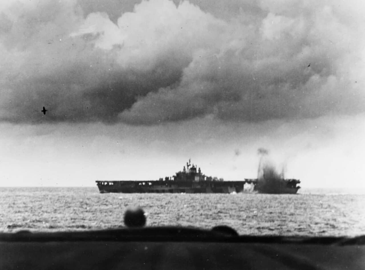 1944年6月19日,「菲律宾海海战」期间:一枚日军炸弹在美军邦克山号航空母舰(CV-17)附近爆炸。画面左侧空中,一架尾部已被击毁的日军战机即将坠毁。注:本照片从「蒙特雷」号轻型航空母舰(CVL-26)上拍摄。